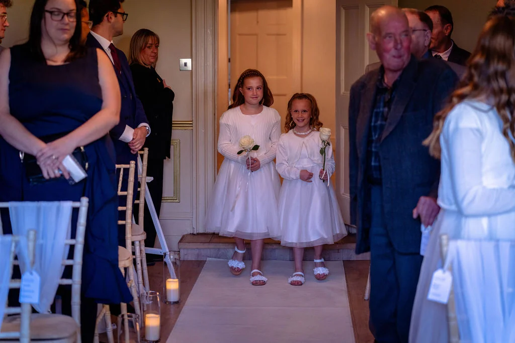 Two young flower girls in white dresses walk down the aisle carrying single white roses as wedding guests look on from chairs adorned with white sashes.