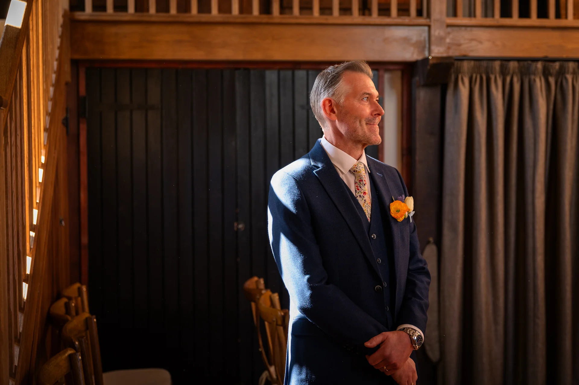 A smiling groom in a navy three-piece suit stands inside a rustic wooden barn, illuminated by soft side light.