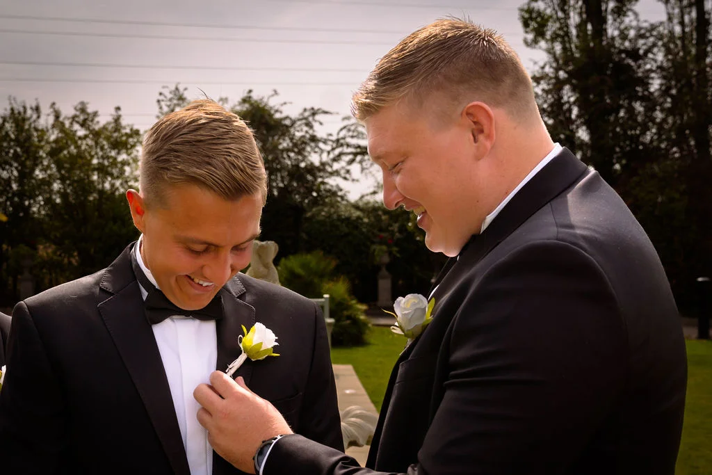 Two grooms in matching black tuxedos and bow ties share a joyful moment outdoors in a garden setting, with one adjusting the other's white rose boutonniere.