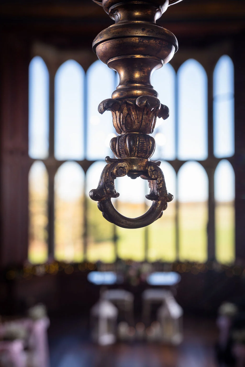 An extreme close-up shot highlights the ornate, gilded brass fixture of a grand hanging light against the softly blurred background of tall arched windows.
