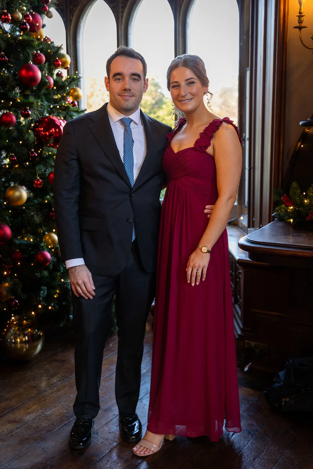 A smiling couple poses indoors next to a large Christmas tree adorned with red and gold baubles, with the man wearing a dark suit and blue tie and the woman wearing a floor-length burgundy gown.