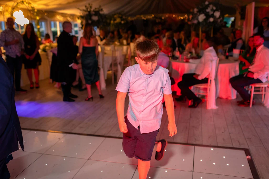 A young guest joyfully dances on an illuminated white dance floor inside a marquee wedding reception illuminated by colourful party lights.