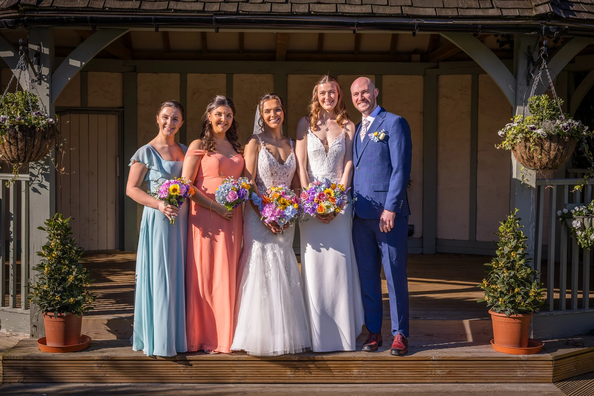 A cheerful wedding party featuring two brides in white gowns, a groom in a blue suit, and two bridesmaids in colorful dresses posing together outdoors on a sunny day.