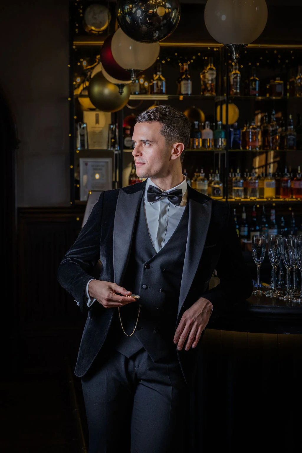 A stylish groom wearing a black velvet tuxedo and waistcoat stands confidently in a dimly lit cocktail bar holding a pocket watch.