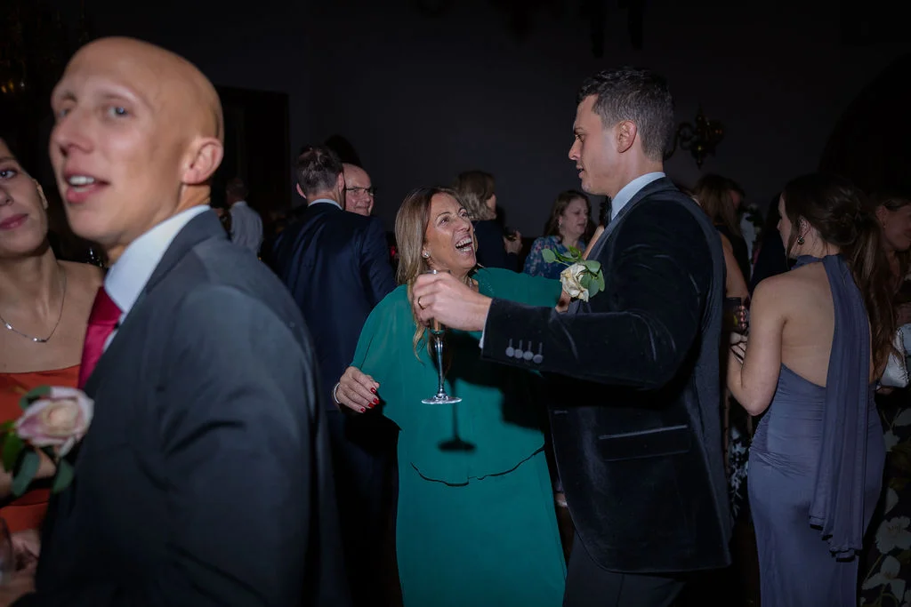 A groom in a velvet tuxedo dances joyfully with a guest holding a champagne glass during the evening wedding reception.