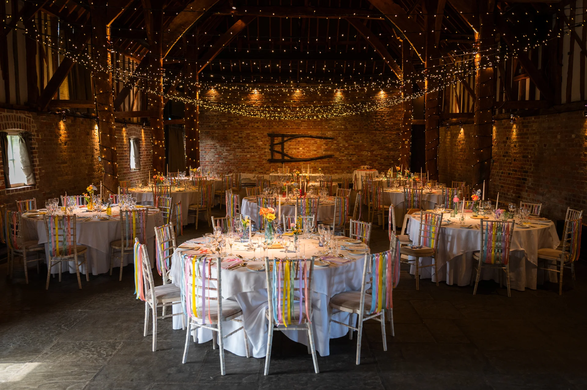 A rustic barn wedding reception featuring round tables adorned with colourful hanging ribbons on chairs beneath a glowing canopy of fairy lights.