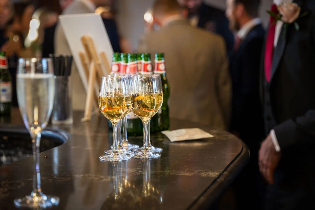 Several wine glasses and a champagne flute sit on a dark bar counter during a wedding drinks reception with guests mingling in the background.