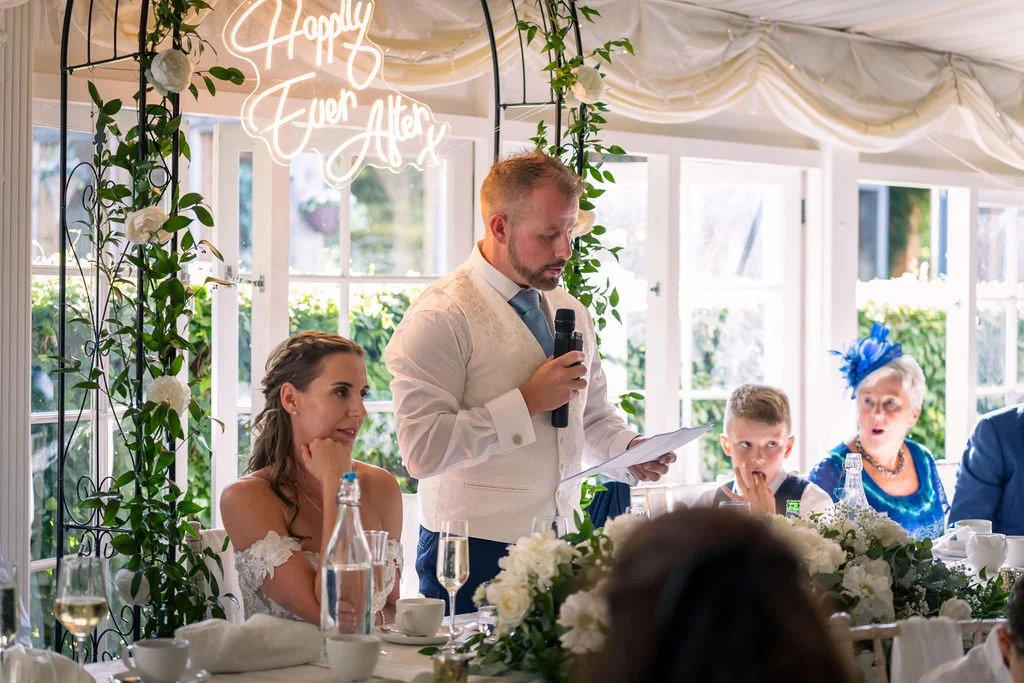 A groom delivers a speech at the top table during a wedding reception, with the bride listening attentively beside him under a floral arch and neon sign.