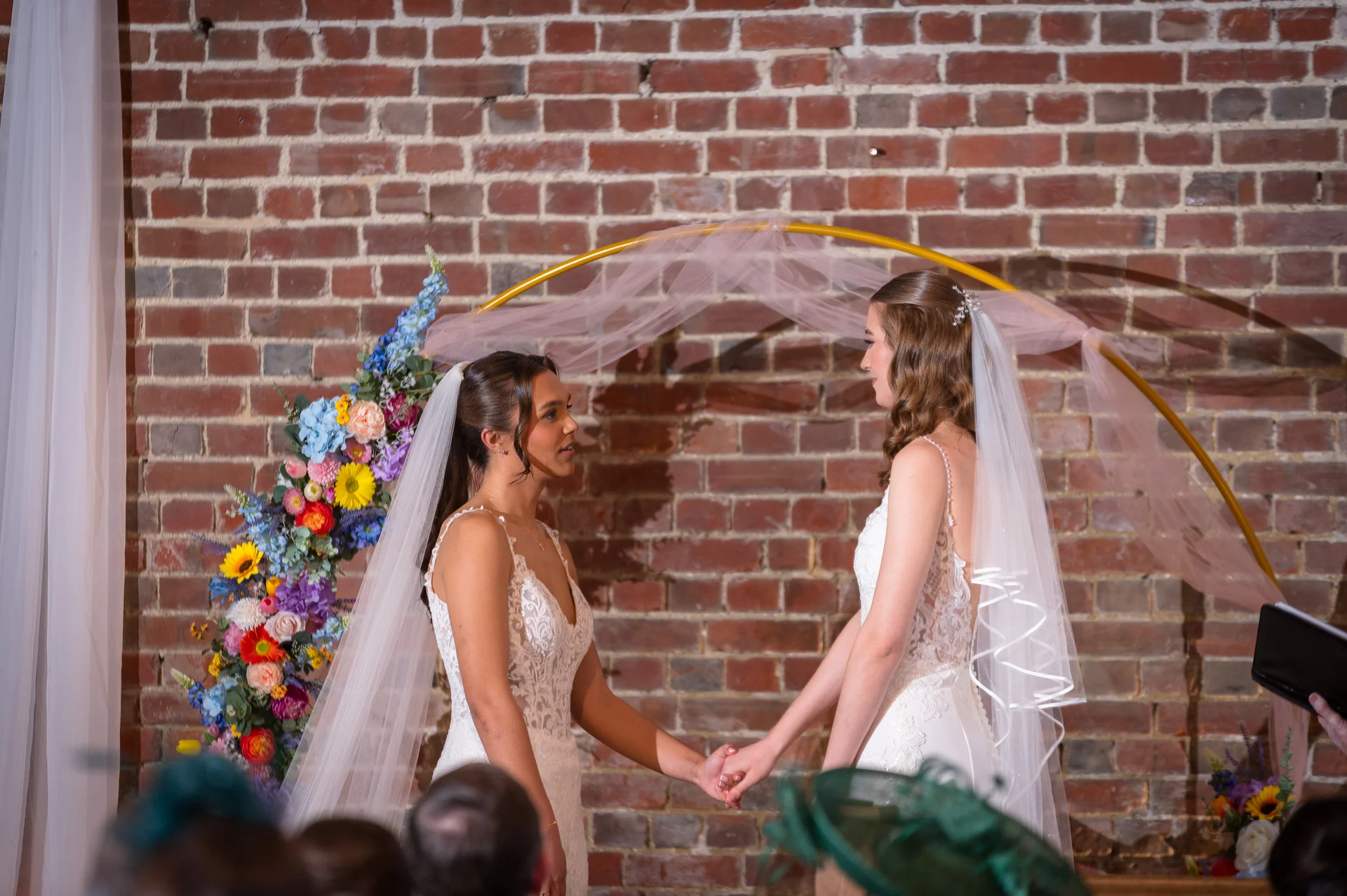 Two brides stand hand in hand during their wedding ceremony in front of an exposed brick wall decorated with a floral circular arch.