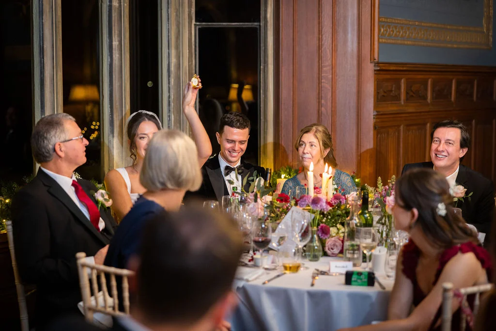 A groom smiling and holding up a pocket watch during speeches at a candlelit wedding breakfast in a grand stately home.