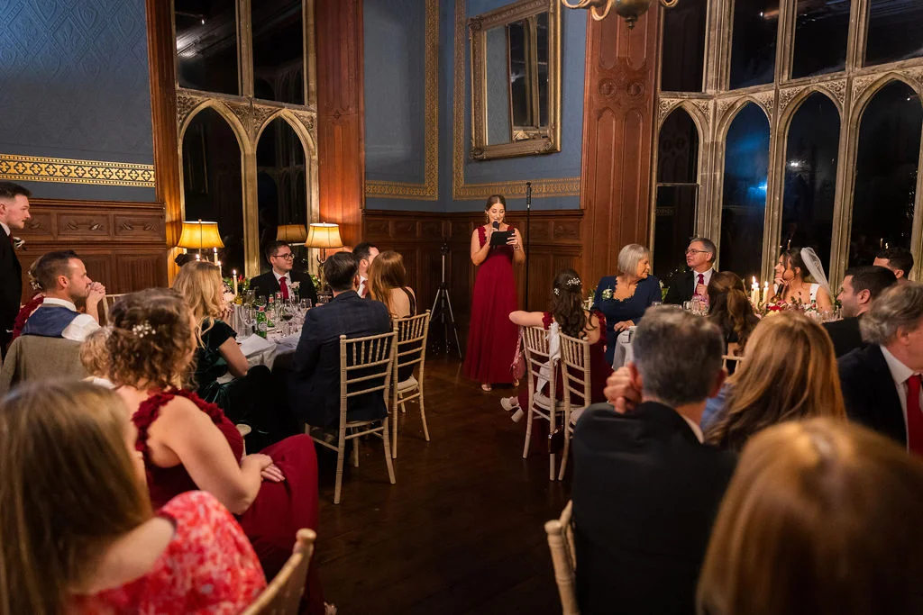 A bridesmaid in a long red dress delivers a speech during the wedding reception in a wood-panelled hall with large arched windows.