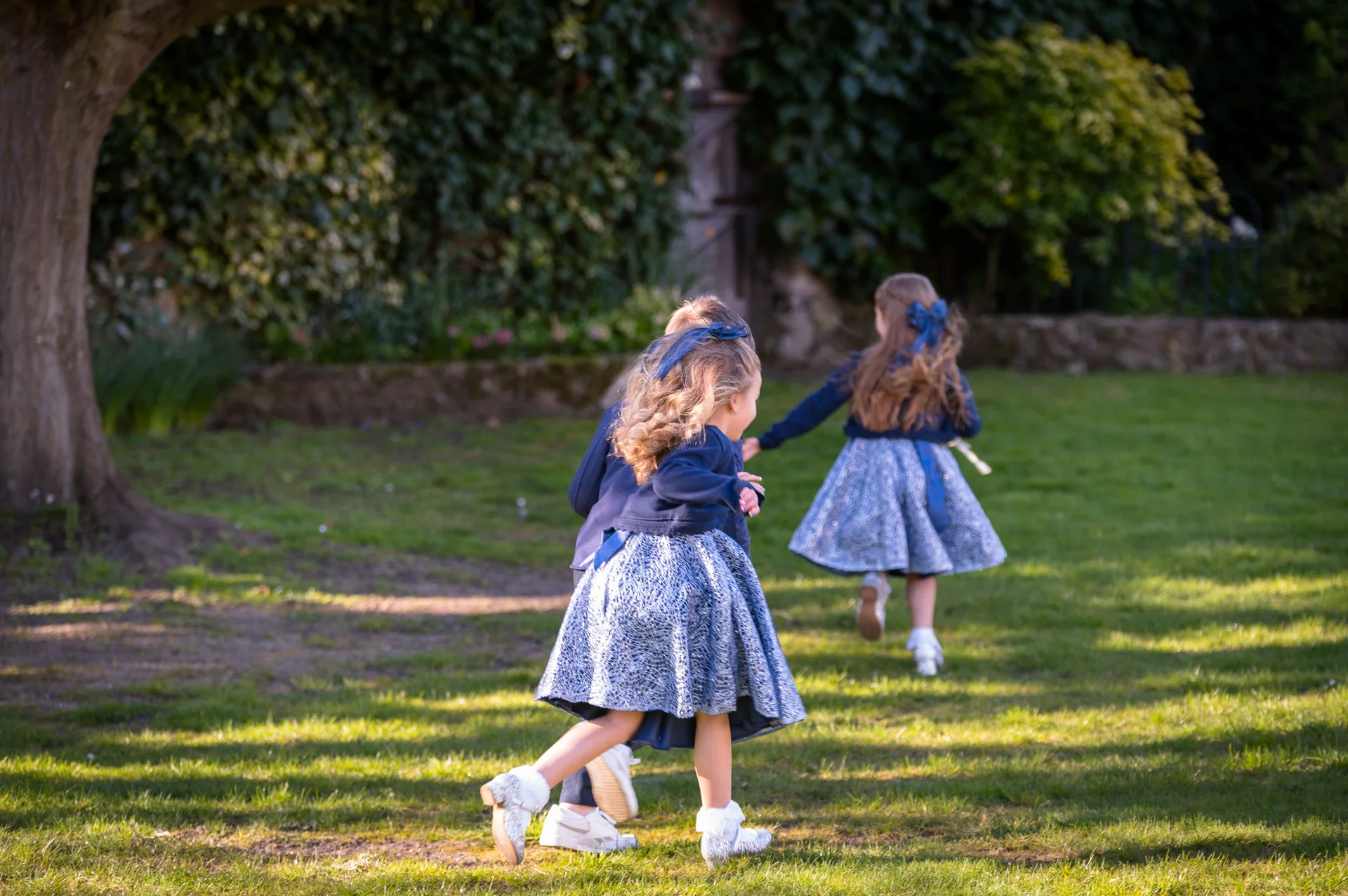 Several young wedding guests dressed in matching blue and silver outfits playfully run across a sunlit manicured lawn.