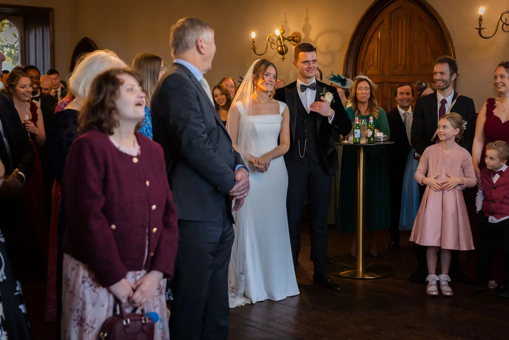The bride in a sleek white gown and groom in a black tuxedo stand among smiling wedding guests inside a historic venue with wooden floors and arched doors.