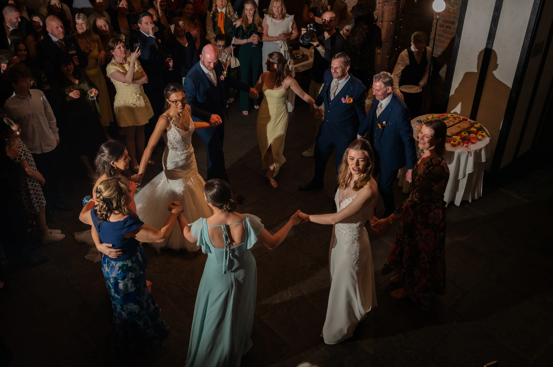 A joyful group of wedding guests and the couple form a large circle on the dance floor, holding hands and laughing during the evening celebration.