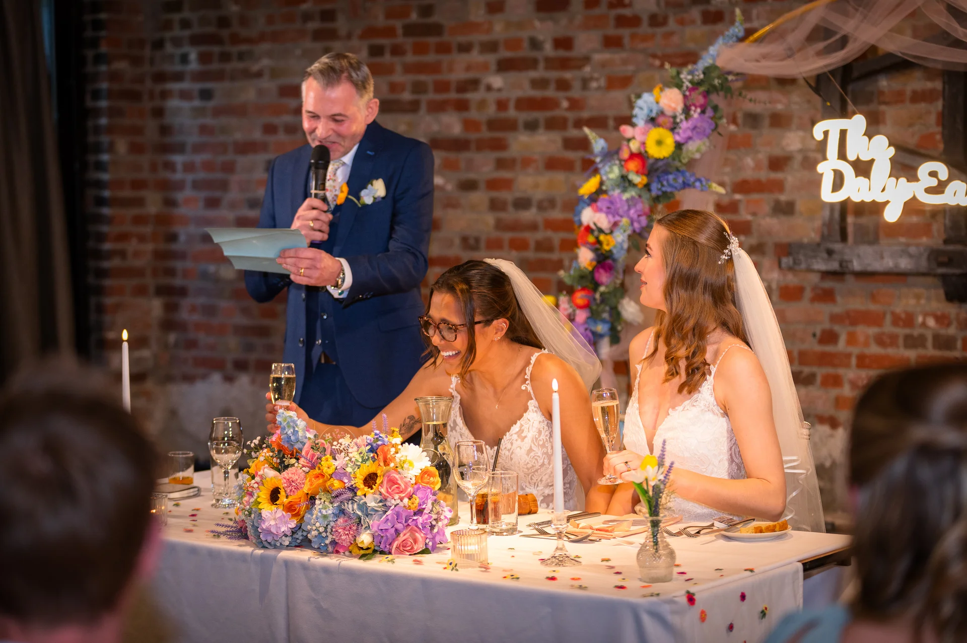 A guest delivers a speech with a microphone as two laughing brides in white wedding dresses hold champagne flutes at a top table decorated with colourful flowers.