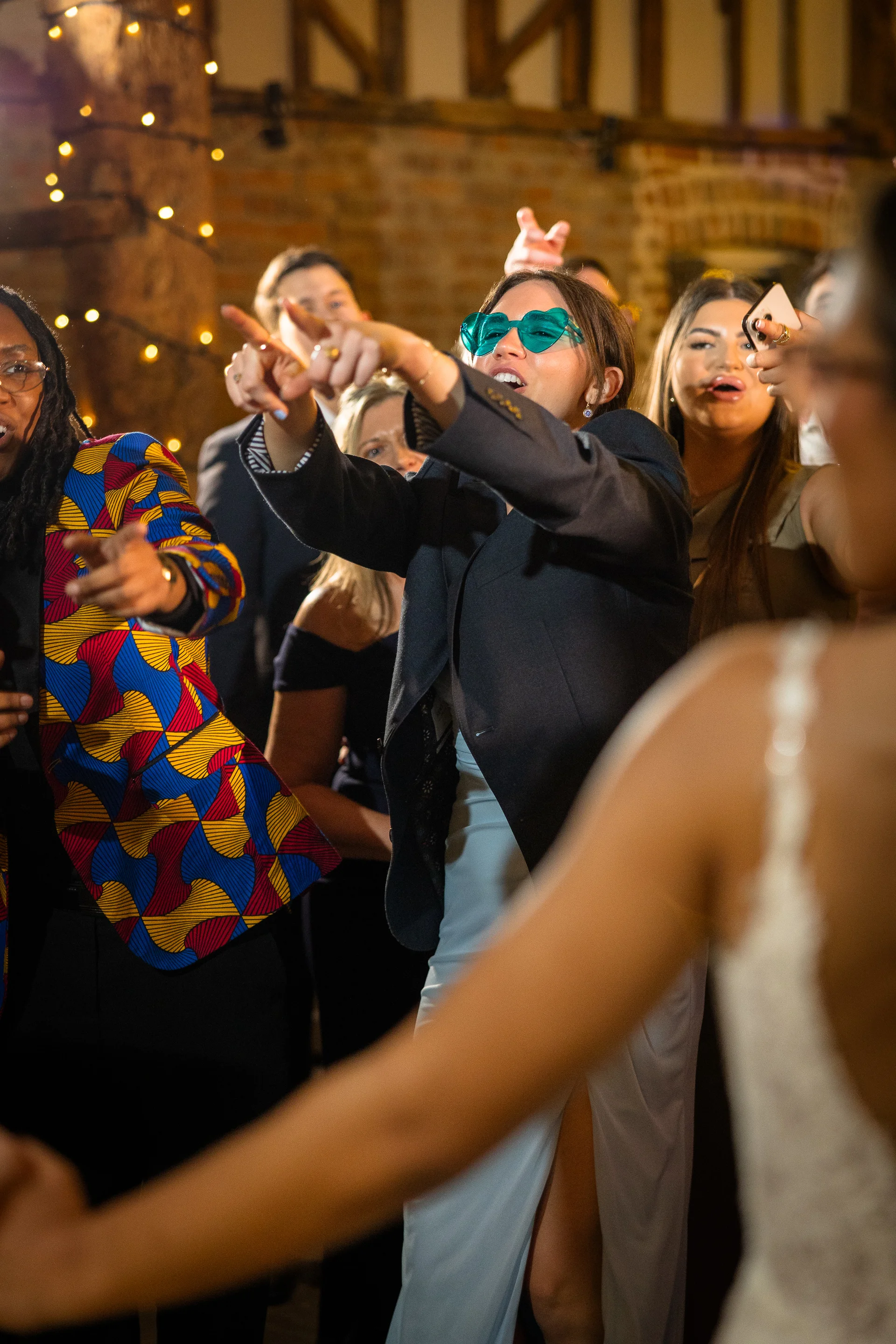A lively group of wedding guests dance and laugh enthusiastically under exposed wooden beams and fairy lights in a rustic barn venue.
