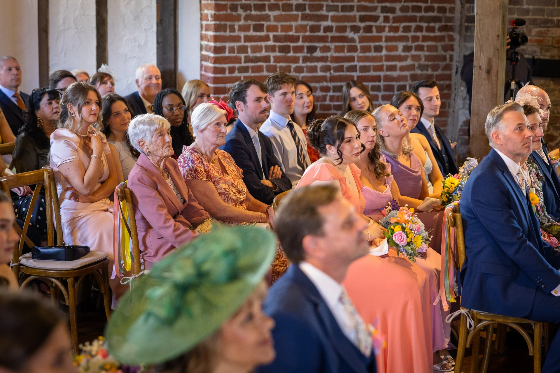 An attentive congregation of well-dressed guests in a rustic brick venue seated on wooden chairs watching a wedding ceremony.