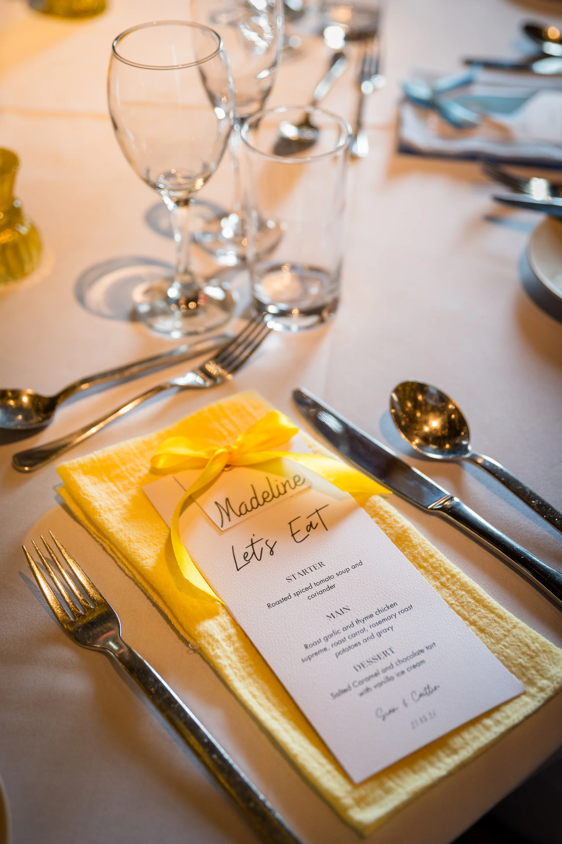 A close up shot of a wedding place setting featuring a yellow napkin, a personalised menu card tied with a yellow ribbon, and polished cutlery.