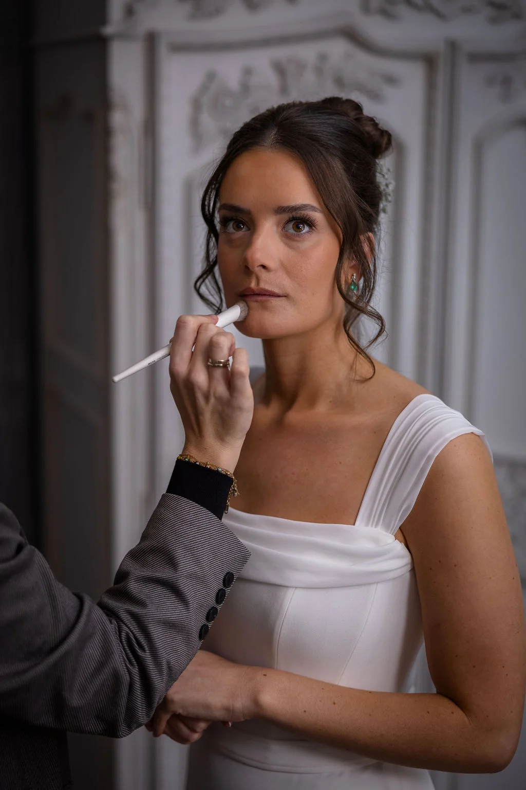 A makeup artist applies finishing touches to a bride with a textured bun and face-framing tendrils while she wears a draped white gown.