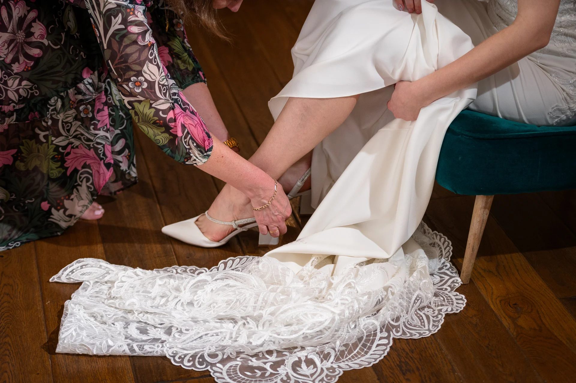 A mother gently helps the bride put on her crystal-embellished satin block heels during bridal preparations.