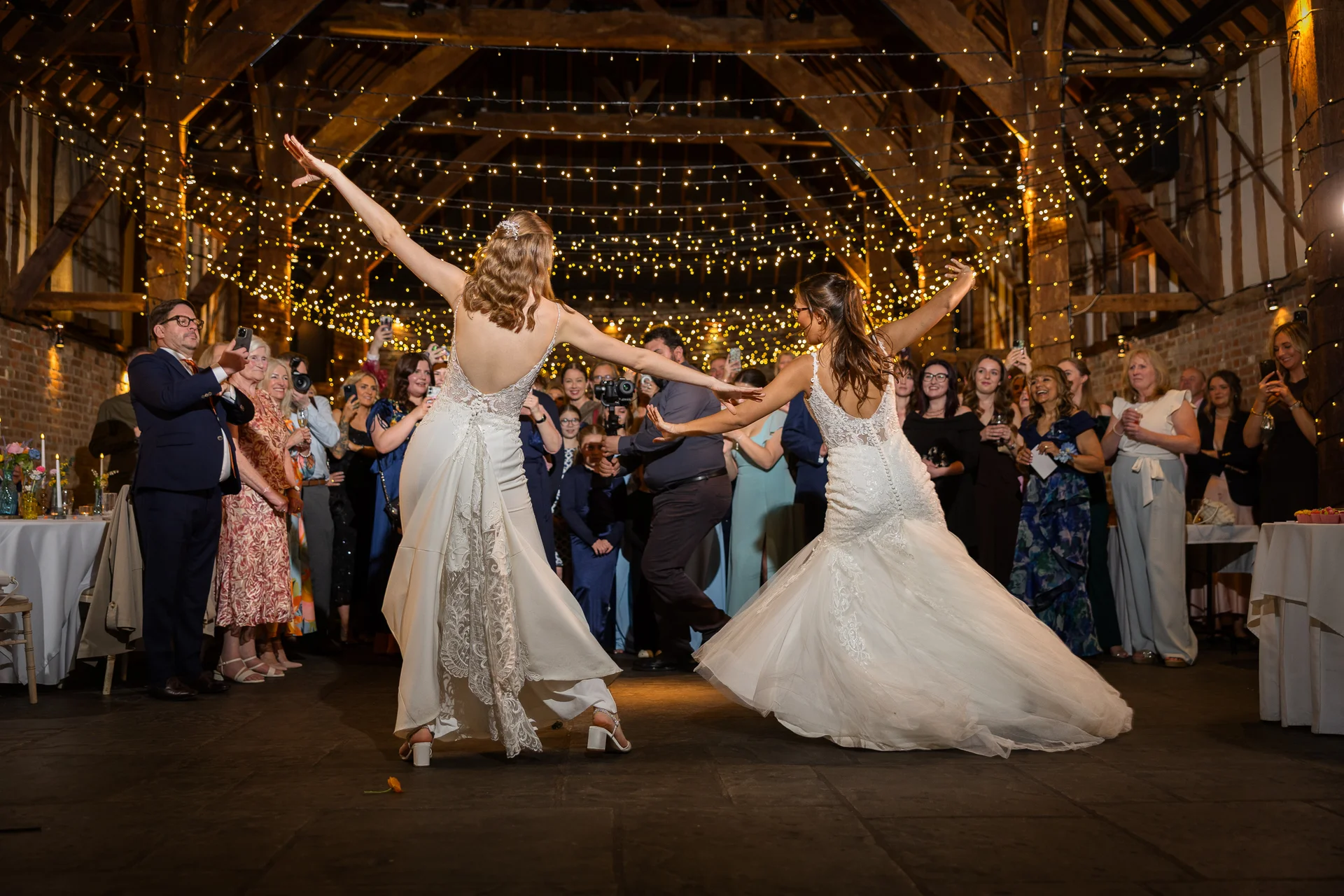 Two brides in white wedding dresses twirl on the dancefloor under a stunning canopy of fairy lights inside a rustic oak-framed barn, surrounded by cheering guests.