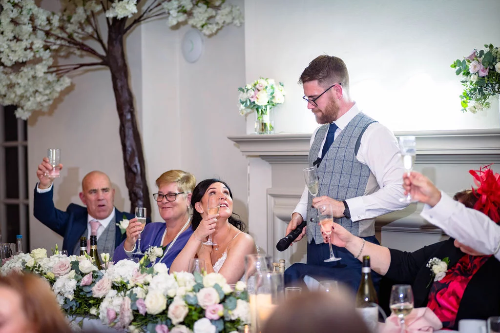 The groom delivers his speech at the top table while the bride looks up admiringly and guests raise their champagne flutes in a toast.