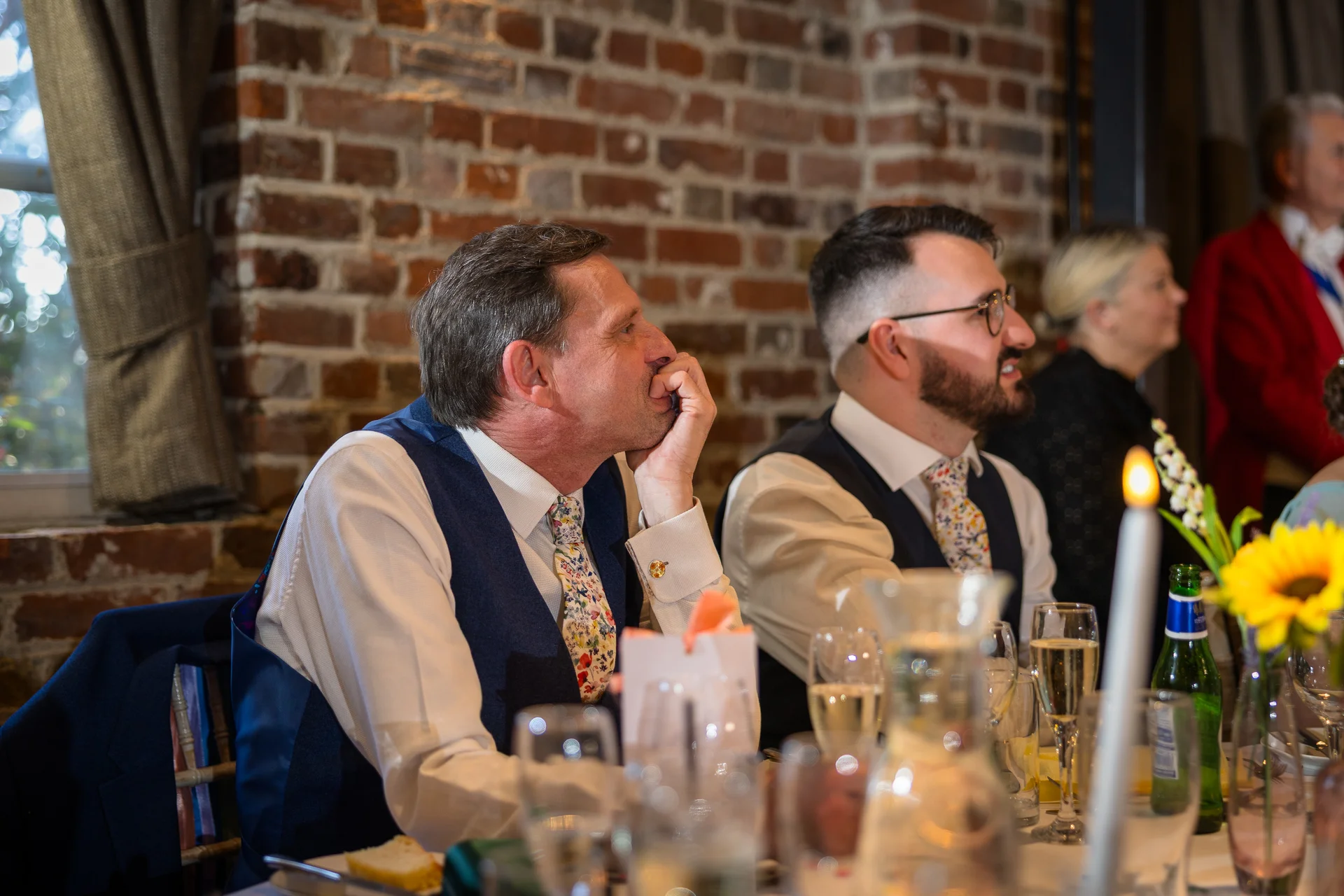 Wedding guests are seated at a long banquet table during the wedding breakfast, laughing while listening to speeches in a venue with exposed brick walls.