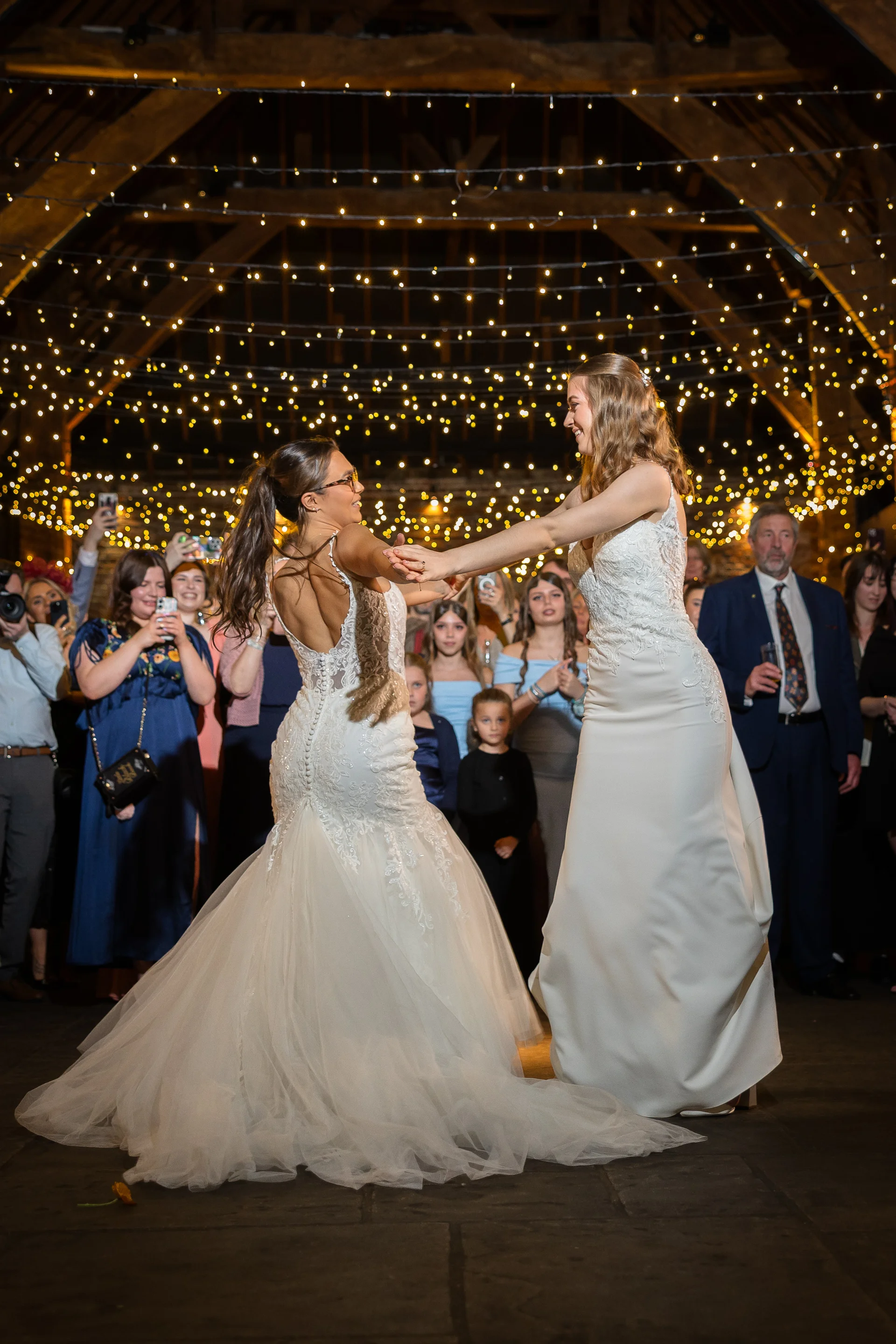 Two brides share their first dance in an oak framed barn illuminated by a stunning overhead fairy light canopy while guests watch and capture the moment.