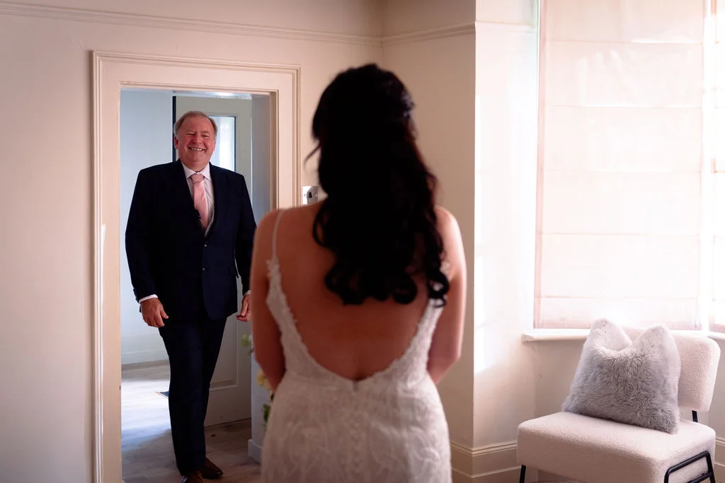 A smiling father in a navy suit and pink tie reacts emotionally during a first look moment with his daughter seen from behind in her wedding dress.