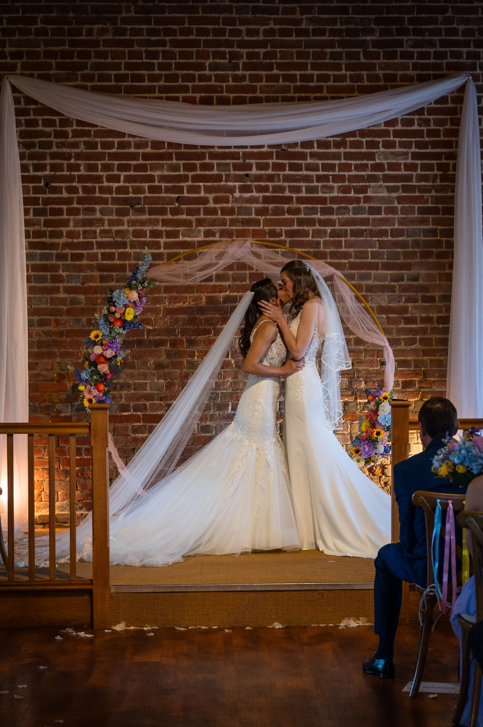 Two brides share a tender first kiss under a floral-adorned circular archway against an exposed brick wall during their indoor ceremony.