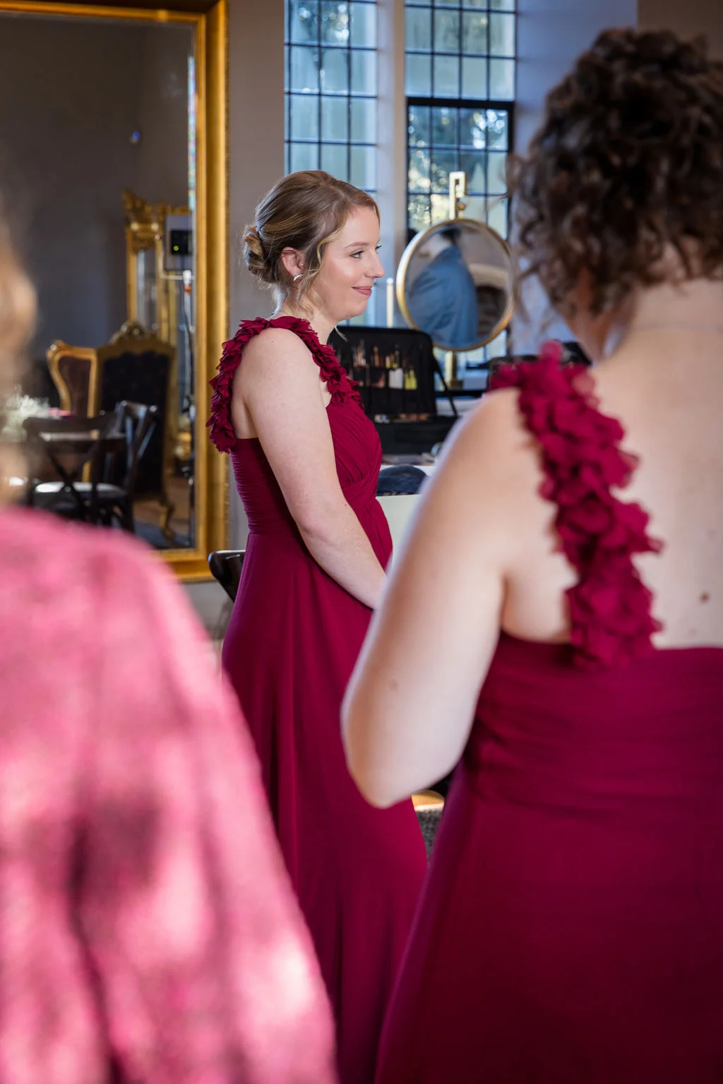 A bridesmaid looks into a mirror while wearing a stunning burgundy dress featuring dramatic ruffled shoulder straps during wedding morning preparations.