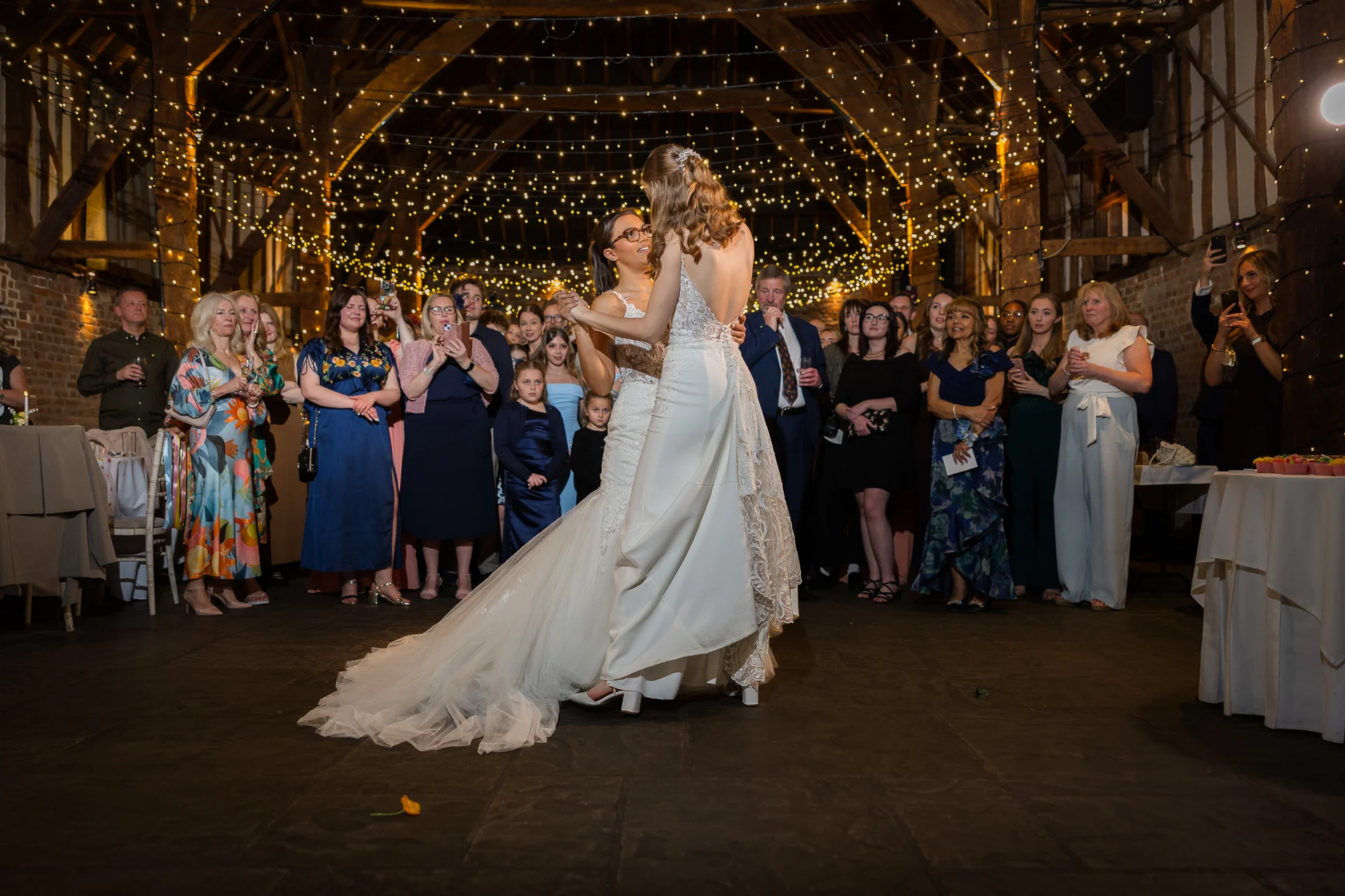 Two brides share their first dance in the centre of a rustic oak-framed barn illuminated by a stunning canopy of twinkling fairy lights while guests watch on.