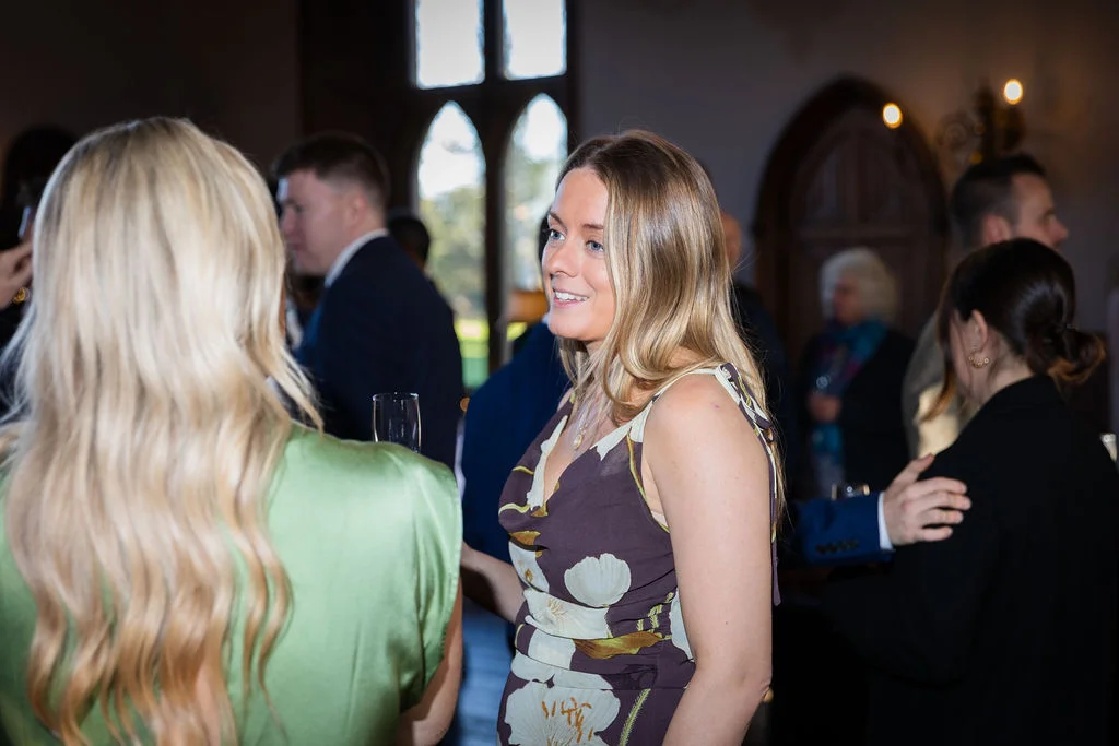 A smiling guest in a floral print dress chats with friends during the drinks reception inside a country house venue.