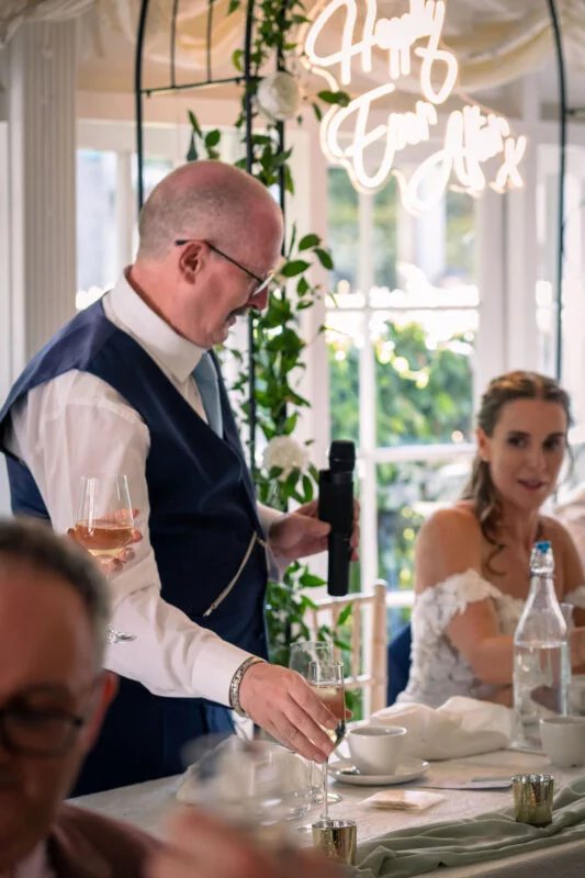 A groom delivers a speech into a microphone at the top table during a wedding reception, with a bride seated nearby and a glowing neon sign hanging in the background.