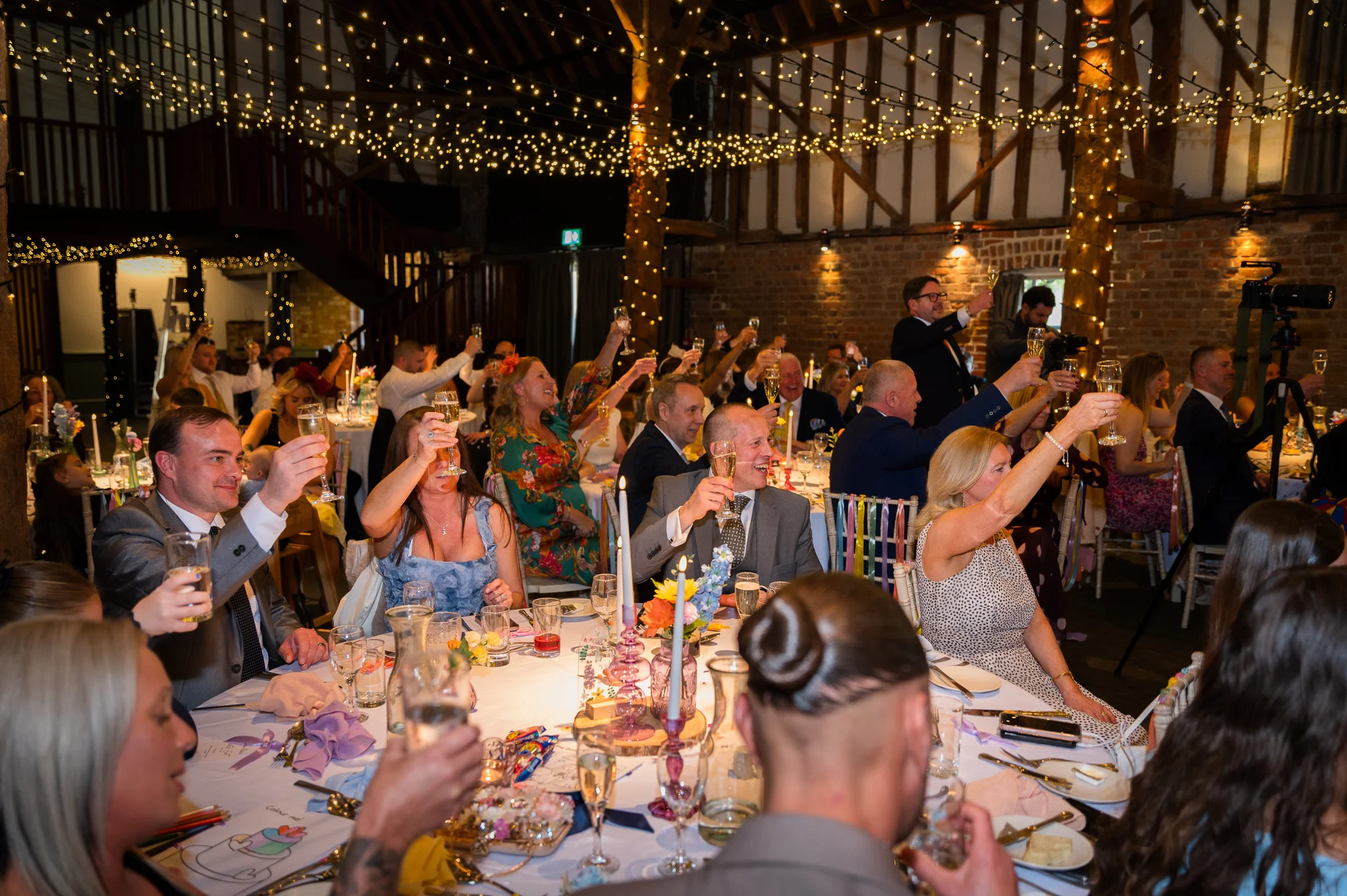 Wedding guests raise their glasses in a joyous toast inside a rustic barn venue illuminated by a canopy of fairy lights.