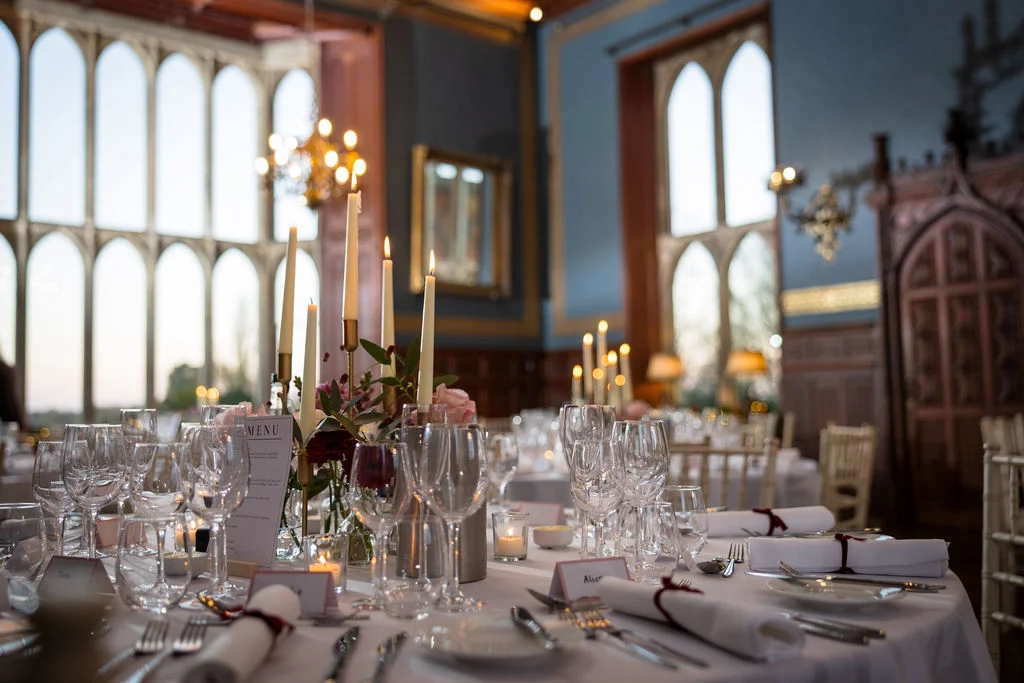 An elegantly dressed round banquet table features tall taper candles in brass holders, crystal glassware, and delicate floral centrepieces inside a grand hall with historic arched windows.