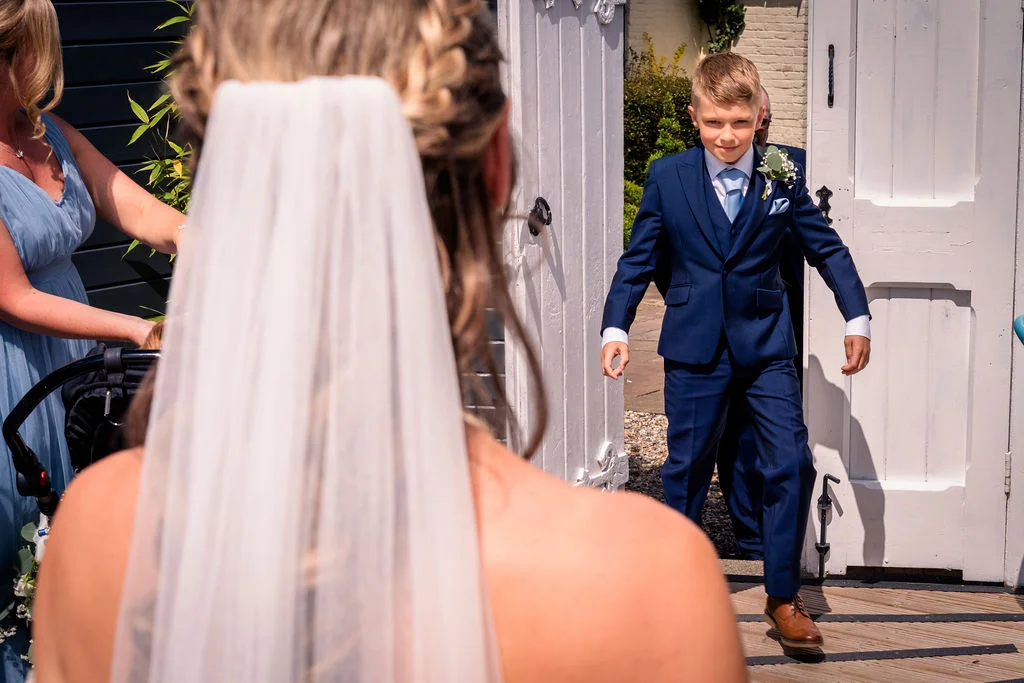 A bride with a braided updo and veil looks over her shoulder as a young page boy in a smart navy suit walks through white wooden doors.