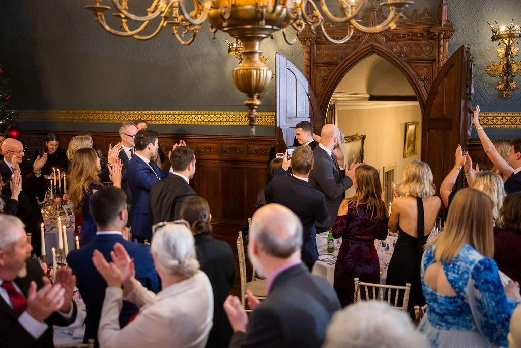 Wedding guests cheer and applaud as the newlyweds make their grand entrance into a historic, wood-paneled banqueting hall lit by glowing candles and a grand chandelier.