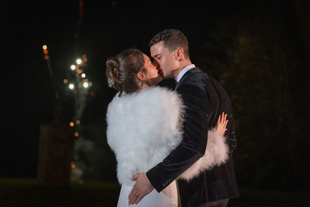 Newlyweds share an intimate kiss outdoors at night, surrounded by a magical bokeh of distant lights while the bride wears a luxurious white faux fur stole.