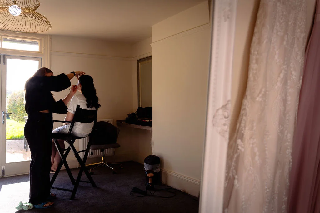 A makeup artist applies finishing touches to a bride sitting in a tall director's chair while her wedding dress hangs in the foreground.