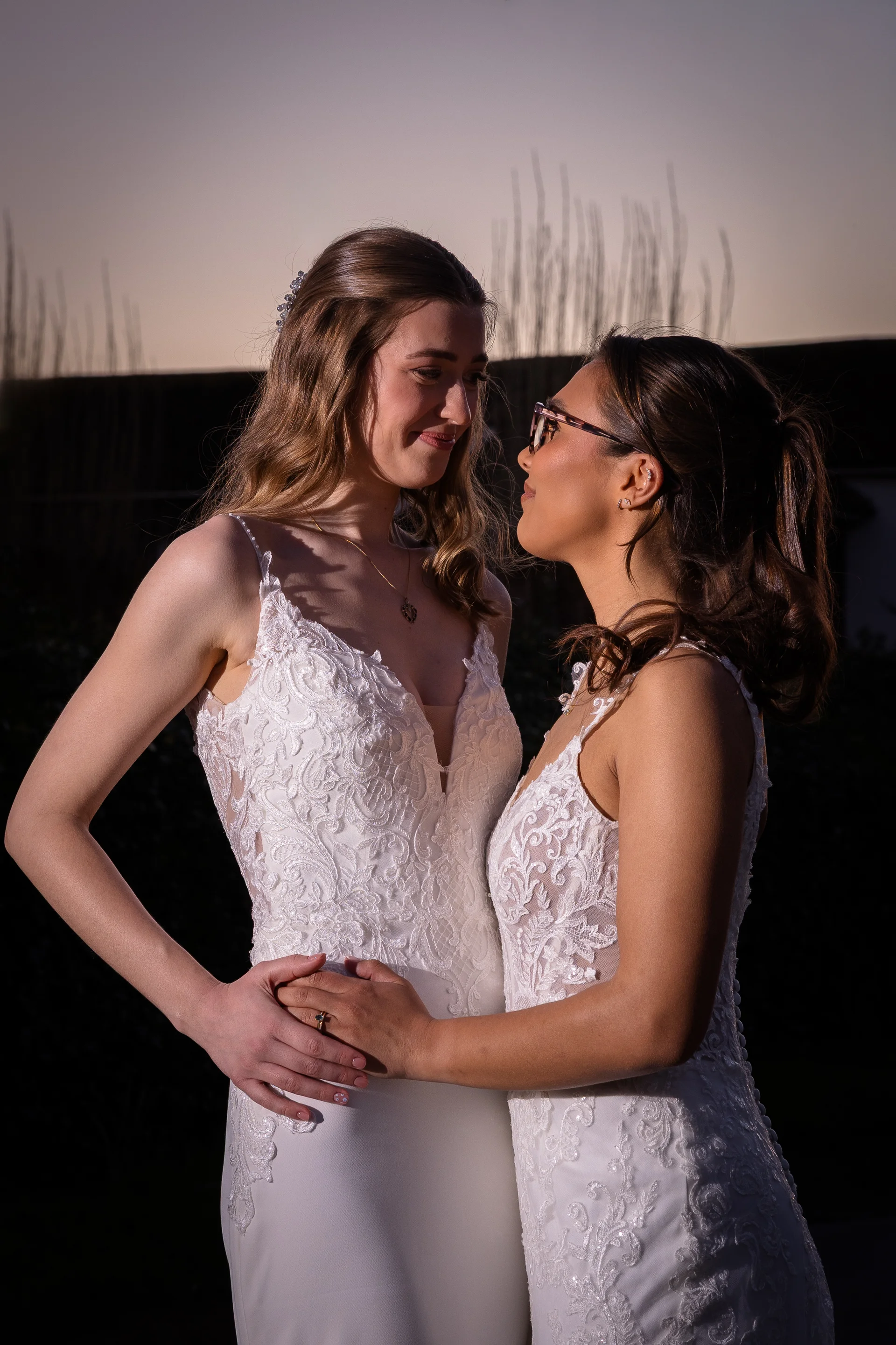 Two smiling brides in lace wedding dresses stand close together outdoors during sunset, gazing affectionately at each other in warm evening light.