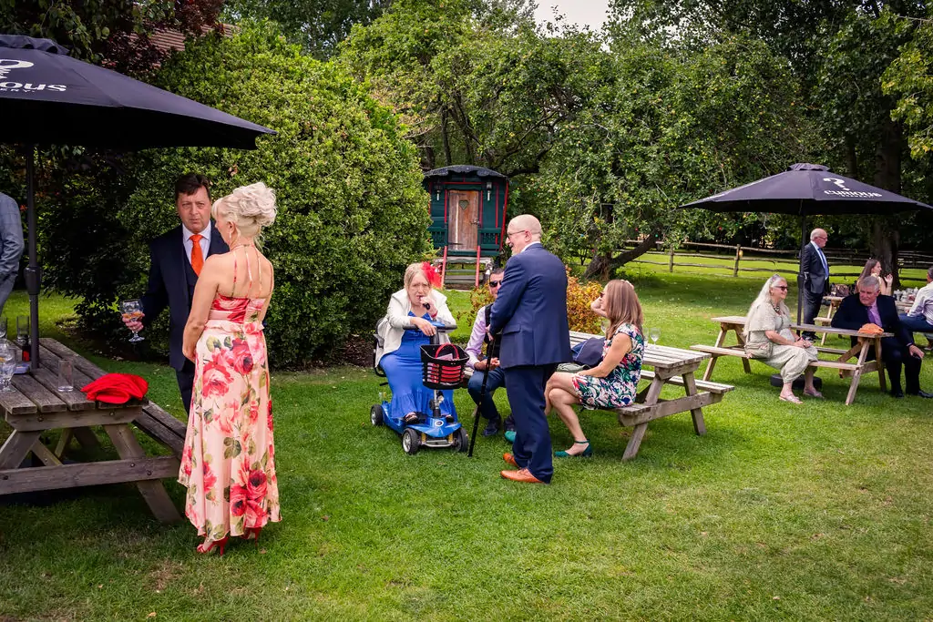 Wedding guests mingle and relax outdoors on the lawn near picnic tables and large black parasols in a lush garden setting.