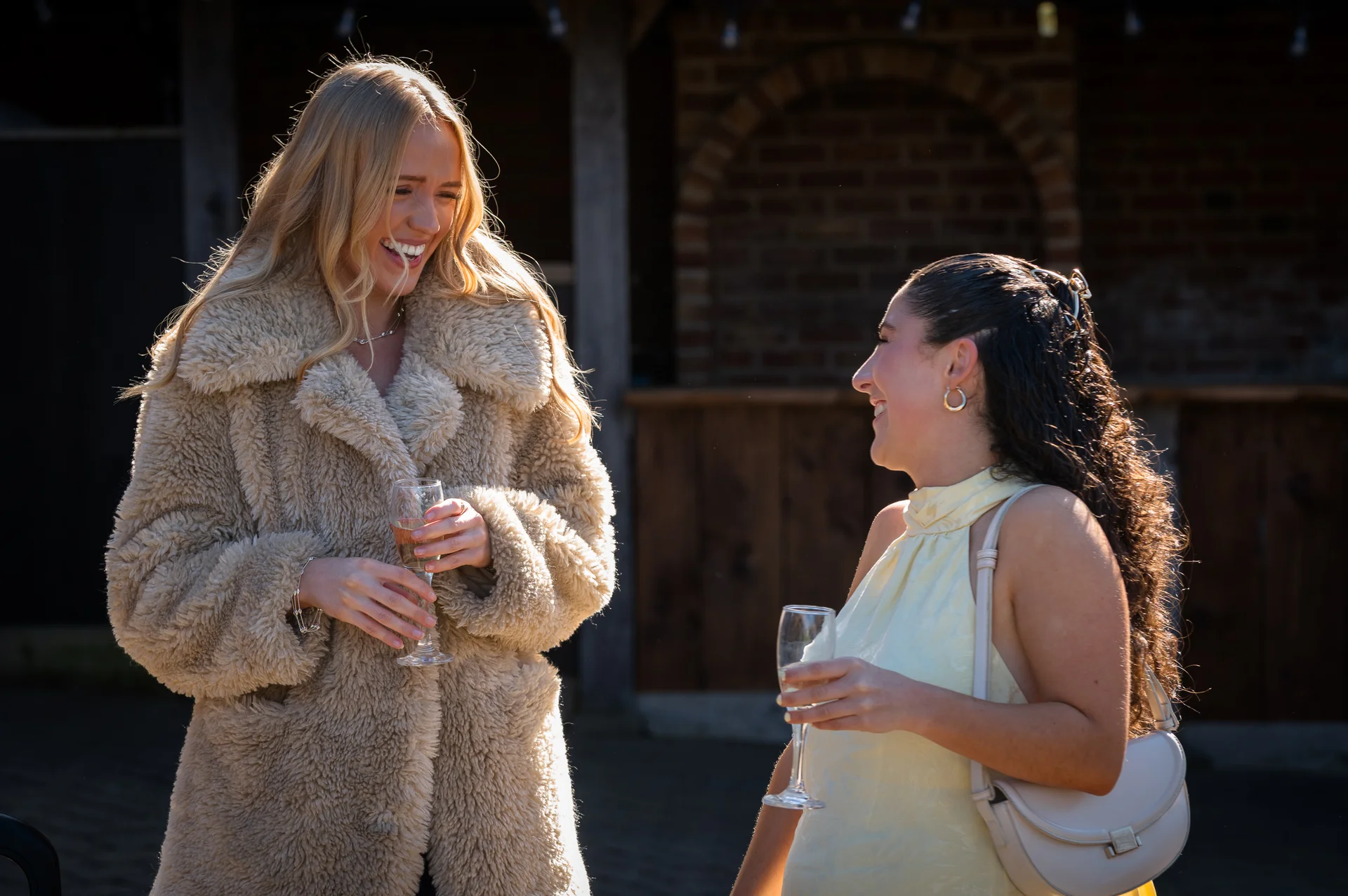 Two wedding guests laugh together in bright sunlight outdoors, holding champagne flutes and enjoying the drinks reception.