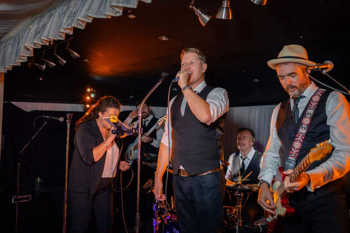 A lively wedding band performs on a stage inside a marquee, with a singer holding a microphone and a guitarist wearing a fedora.