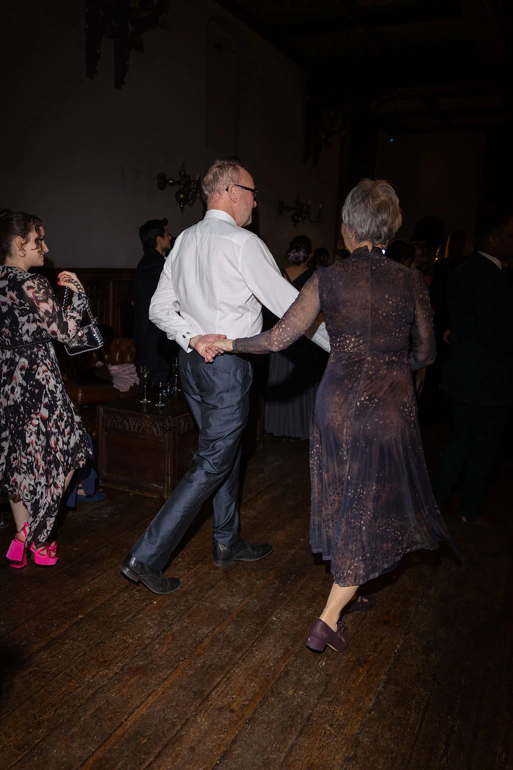 An older couple enjoys dancing together on a wooden floor at a dimly lit evening wedding reception.