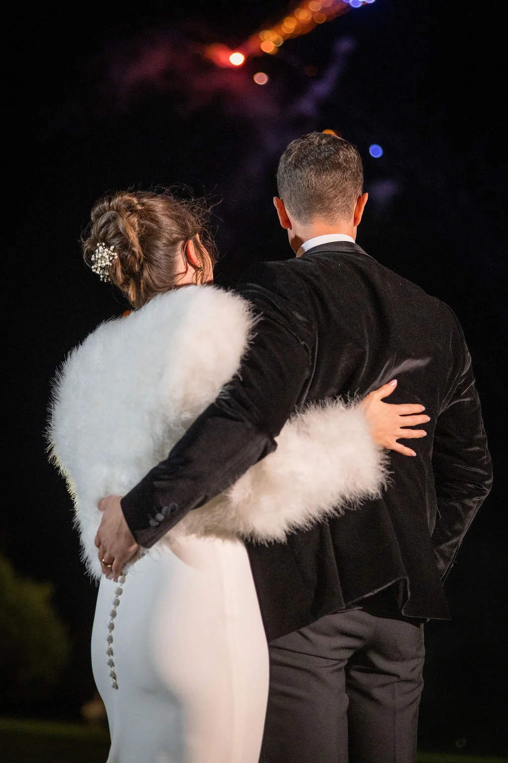 A newlywed couple viewed from behind embraces tightly while watching a dramatic fireworks display light up the night sky.