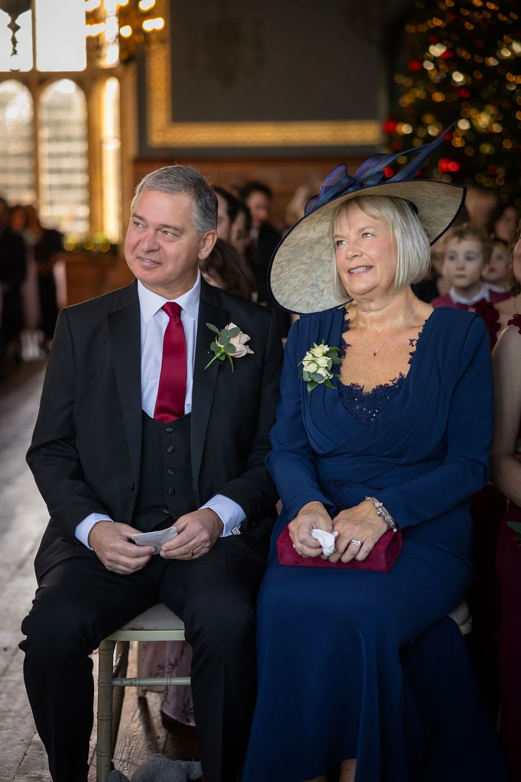 An elegantly dressed older couple, the mother and father of the bride, sit together smiling during a wedding ceremony inside a historic venue.