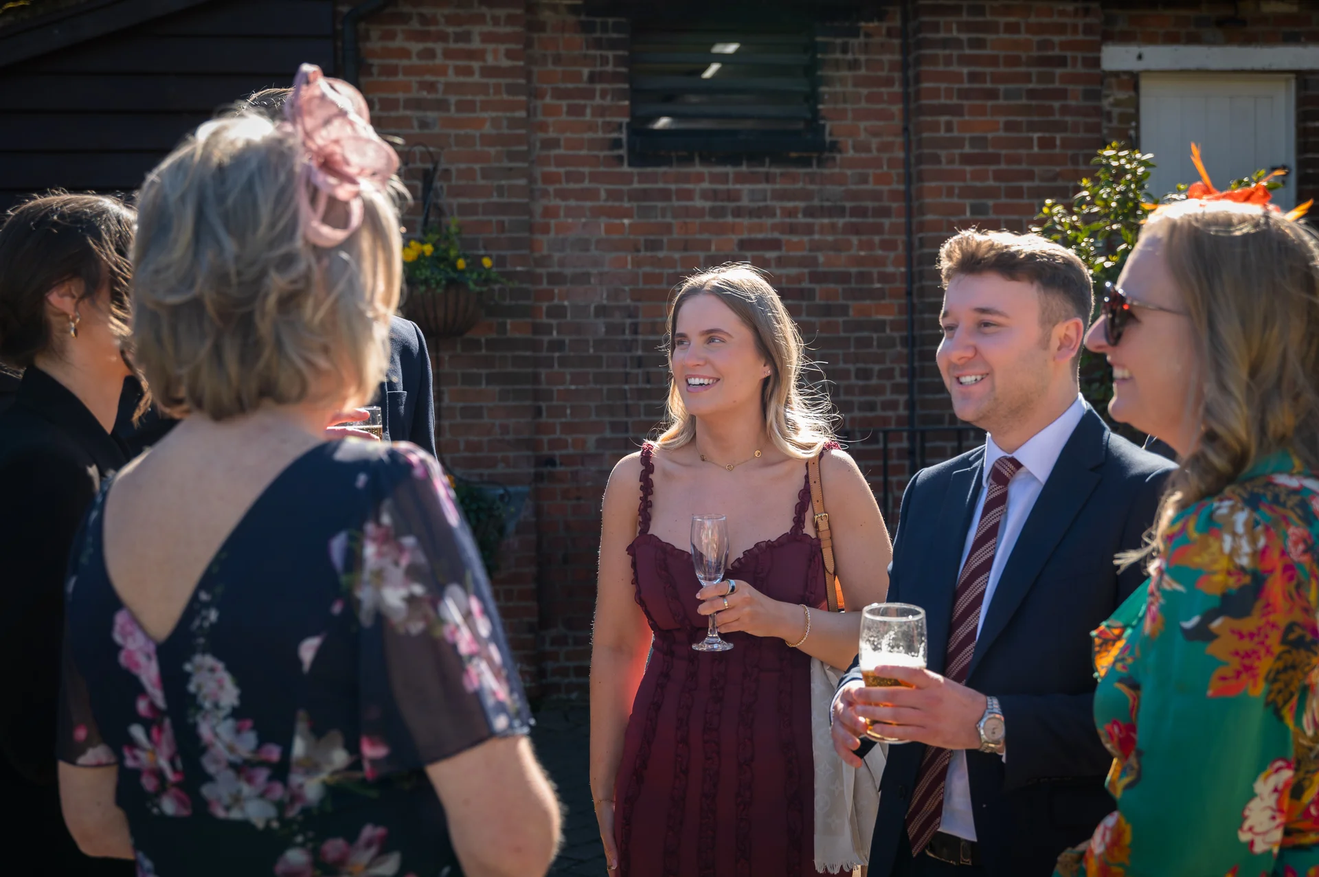 Wedding guests enjoy chatting and drinks in the sunshine outdoors against a rustic brick wall during the reception.