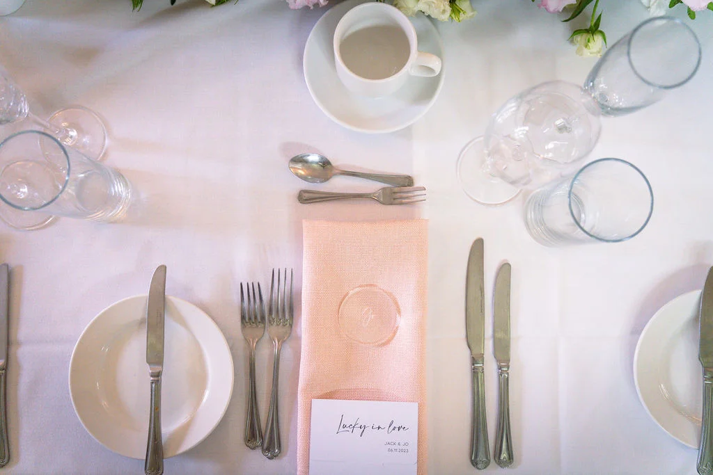An overhead flat lay view of a wedding tablescape featuring a peach napkin, minimalist place card, white plates, and gleaming cutlery on a crisp white tablecloth.