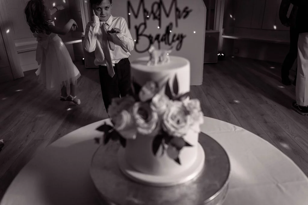 A two-tier wedding cake adorned with floral arrangements sits in the foreground with children dancing on the reception dance floor behind it.